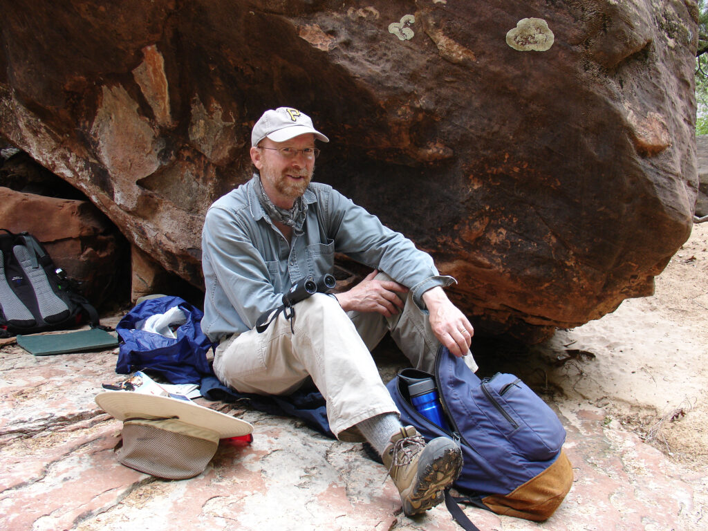 Pat McShea sitting next to a boulder with binoculars on his lap and hiking gear (backpack, hat, jacket) scattered around him.
